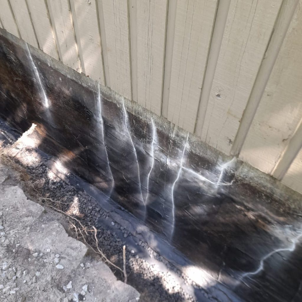 Foundation wall coated in black waterproofing with sealed vertical cracks above a gravel trench